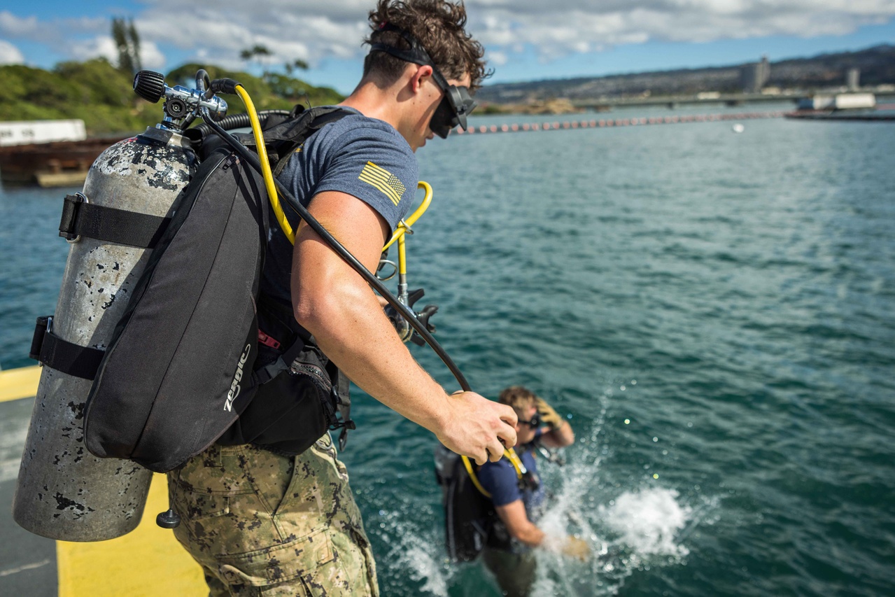 Two people in scuba-diving equipment jump into the water.