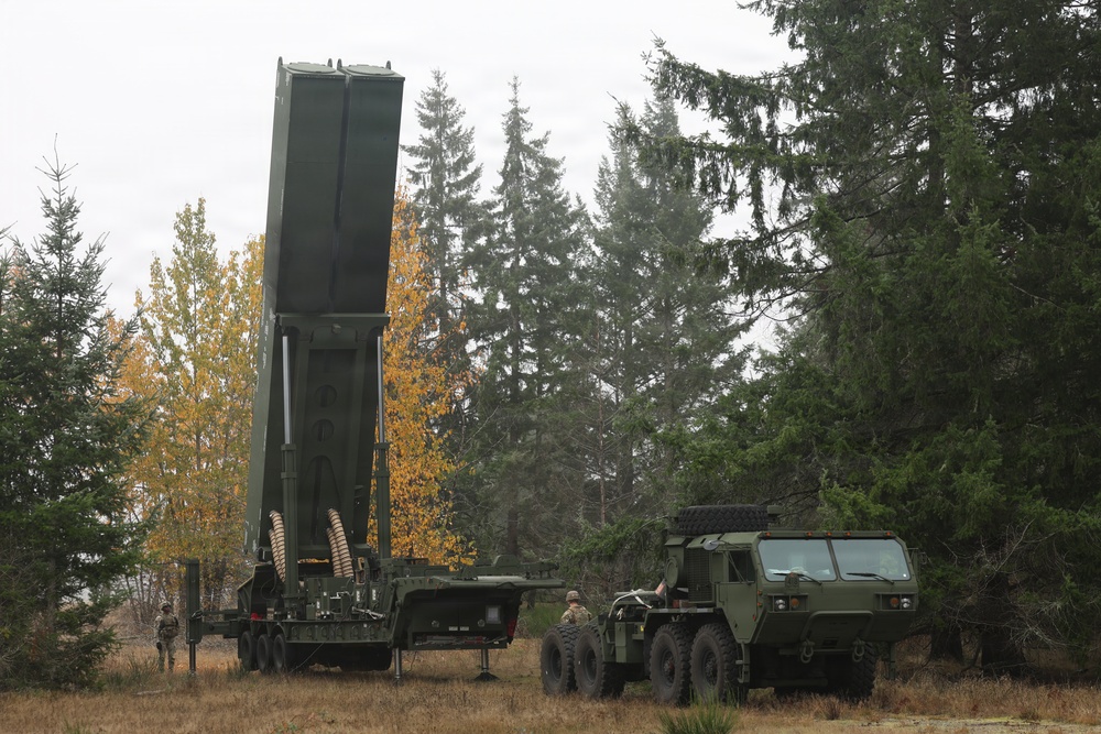 The image shows a military missile launcher system, consistent with a HIMARS or similar large rocket artillery platform, deployed in a forested clearing with autumn foliage visible. The launcher pod is elevated to a near-vertical position, suggesting a setup or training exercise. Several military personnel and a heavy tactical truck are present alongside the equipment.