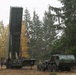 The image shows a military missile launcher system, consistent with a HIMARS or similar large rocket artillery platform, deployed in a forested clearing with autumn foliage visible. The launcher pod is elevated to a near-vertical position, suggesting a setup or training exercise. Several military personnel and a heavy tactical truck are present alongside the equipment.