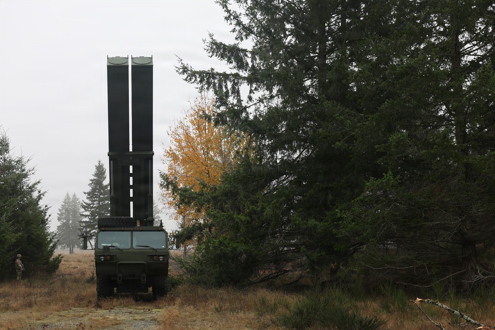A military vehicle with a large vertically-oriented launcher or antenna system is positioned in a forested area during overcast autumn conditions. The vehicle appears to be a tactical military truck with a tall dual-tube structure mounted on its bed, likely a missile launcher or radar system. At least one soldier in uniform is visible in the background to the left.