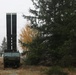 A military vehicle with a large vertically-oriented launcher or antenna system is positioned in a forested area during overcast autumn conditions. The vehicle appears to be a tactical military truck with a tall dual-tube structure mounted on its bed, likely a missile launcher or radar system. At least one soldier in uniform is visible in the background to the left.