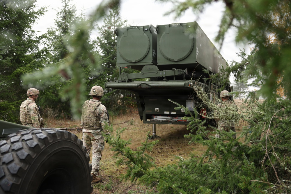 Three soldiers in camouflage uniforms and helmets are observing a large military missile launcher system positioned in a wooded area. The launcher appears to be a trailer-mounted system with two large circular launch tubes, consistent with a surface-to-air missile system. The equipment is positioned among conifer trees, suggesting a concealed or field deployment scenario.