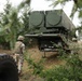 Three soldiers in camouflage uniforms and helmets are observing a large military missile launcher system positioned in a wooded area. The launcher appears to be a trailer-mounted system with two large circular launch tubes, consistent with a surface-to-air missile system. The equipment is positioned among conifer trees, suggesting a concealed or field deployment scenario.