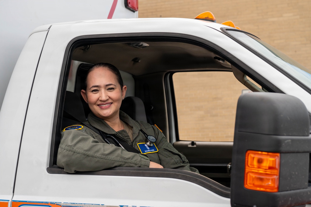 An airman sitting in the passenger seat of an ambulance smiles and looks out of an open window during daytime, with a building in the background.