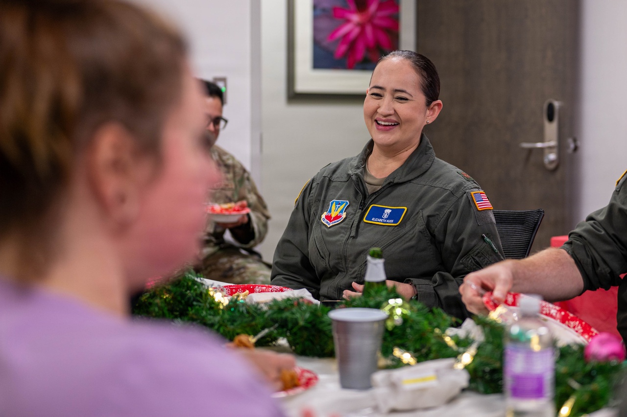 An airman in a flight suit sits and smiles at a table with food on paper plates, green decorations and other people indoors.