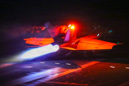 A jet lit with red/orange light takes off at night from the deck of a ship with light blue flame shooting out from the back.
