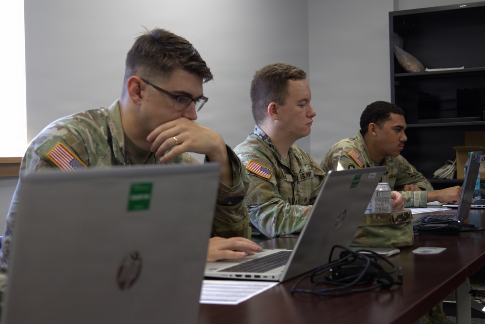Three U.S. Army soldiers in MultiCam/OCP camouflage uniforms are seated at a table working on laptops in what appears to be an office or classroom setting. All three wear American flag patches on their right shoulders, indicating active U.S. Army personnel. They appear to be engaged in a training session, briefing, or administrative work.