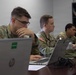 Three U.S. Army soldiers in MultiCam/OCP camouflage uniforms are seated at a table working on laptops in what appears to be an office or classroom setting. All three wear American flag patches on their right shoulders, indicating active U.S. Army personnel. They appear to be engaged in a training session, briefing, or administrative work.