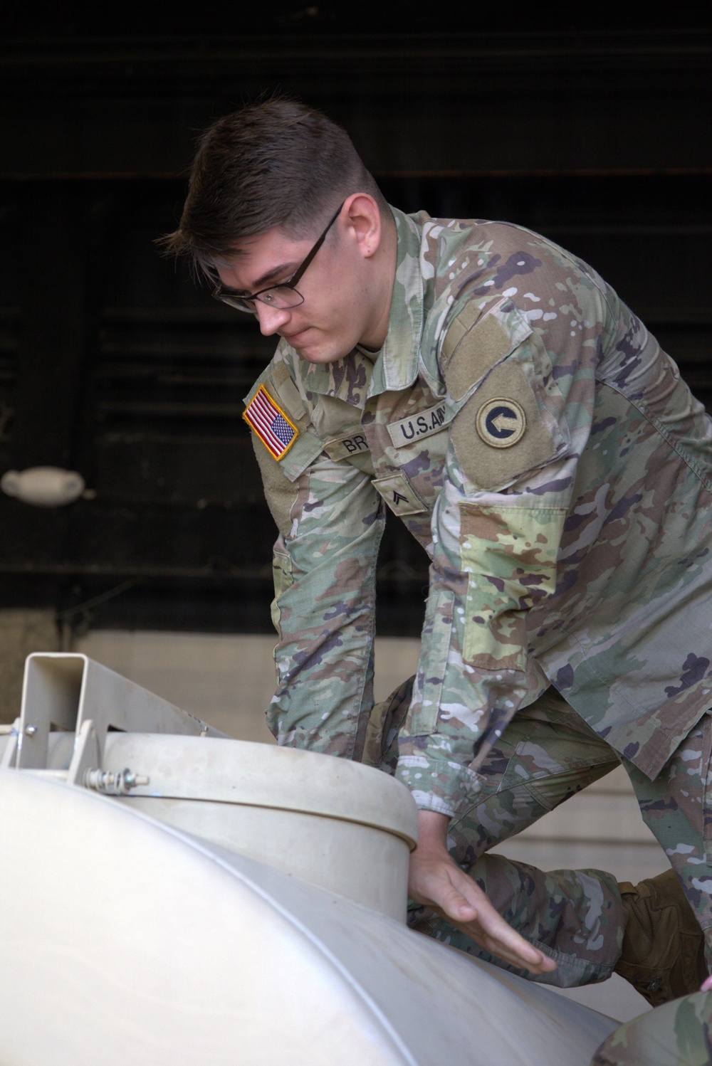 A U.S. Army soldier in Operational Camouflage Pattern (OCP) uniform is working on a large white cylindrical or dome-shaped piece of equipment. The soldier's name tape begins with 'BR' and rank insignia indicates Private First Class. The setting appears to be a maintenance bay or hangar environment.