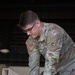 A U.S. Army soldier in Operational Camouflage Pattern (OCP) uniform is working on a large white cylindrical or dome-shaped piece of equipment. The soldier's name tape begins with 'BR' and rank insignia indicates Private First Class. The setting appears to be a maintenance bay or hangar environment.