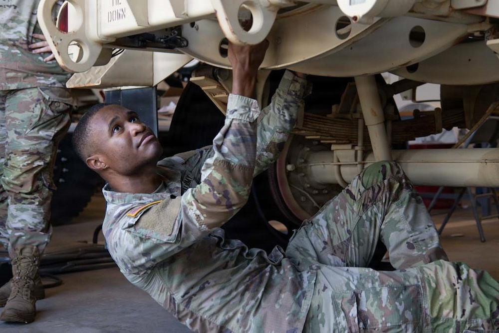A U.S. Army soldier in Operational Camouflage Pattern (OCP) uniform is lying on their back performing maintenance work on the underside of a military vehicle. A second soldier in the same uniform is visible standing nearby, assisting with the task. The work appears to be taking place inside a maintenance bay or hangar facility.