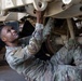 A U.S. Army soldier in Operational Camouflage Pattern (OCP) uniform is lying on their back performing maintenance work on the underside of a military vehicle. A second soldier in the same uniform is visible standing nearby, assisting with the task. The work appears to be taking place inside a maintenance bay or hangar facility.