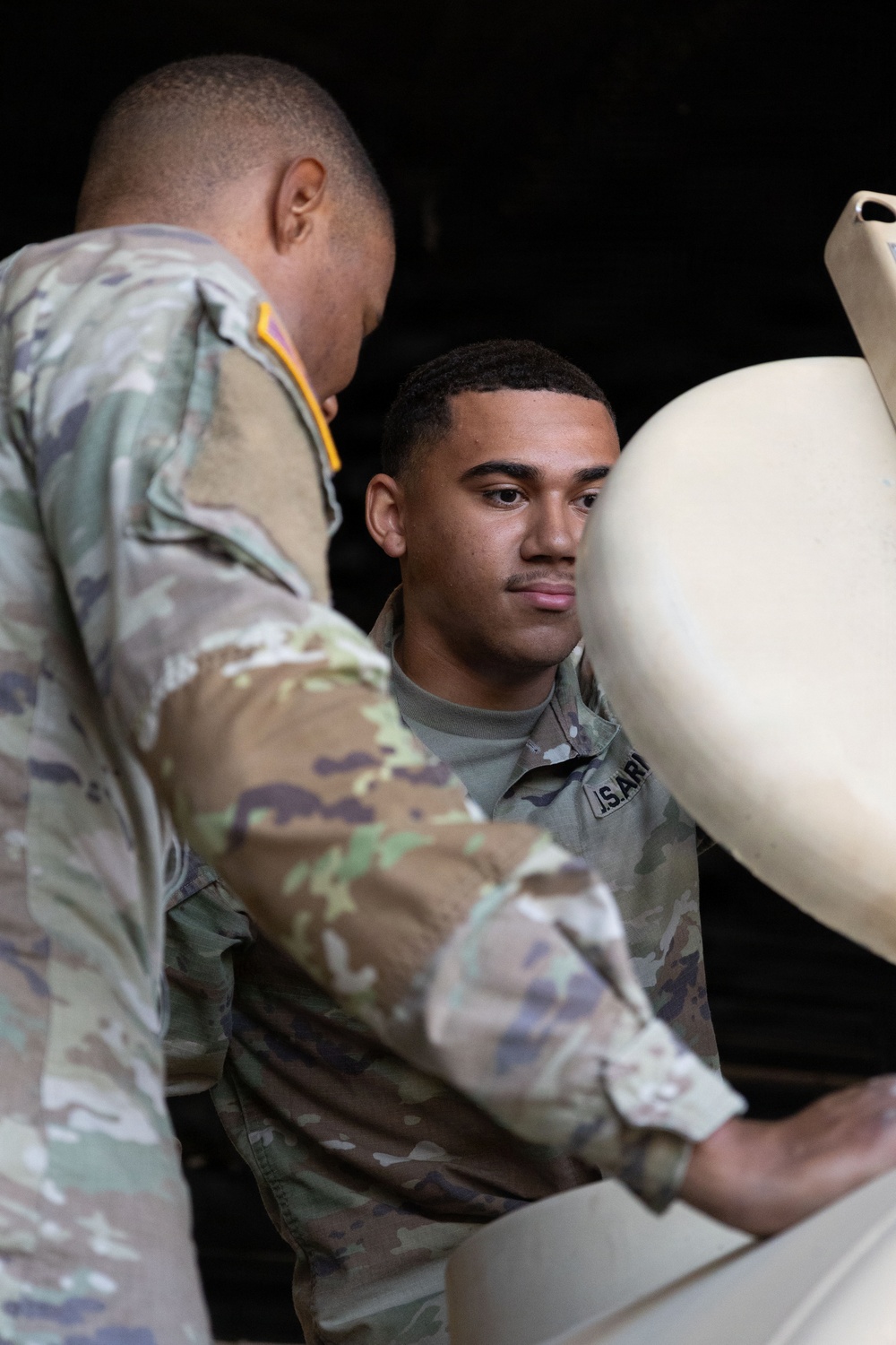 Two U.S. Army soldiers in OCP (Operational Camouflage Pattern) uniforms are handling a large tan/beige military container or equipment piece. The soldier facing the camera has a 'U.S. ARMY' tape visible on his uniform. They appear to be loading, unloading, or inspecting the item in what appears to be a dimly lit storage or vehicle area.