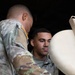 Two U.S. Army soldiers in OCP (Operational Camouflage Pattern) uniforms are handling a large tan/beige military container or equipment piece. The soldier facing the camera has a 'U.S. ARMY' tape visible on his uniform. They appear to be loading, unloading, or inspecting the item in what appears to be a dimly lit storage or vehicle area.