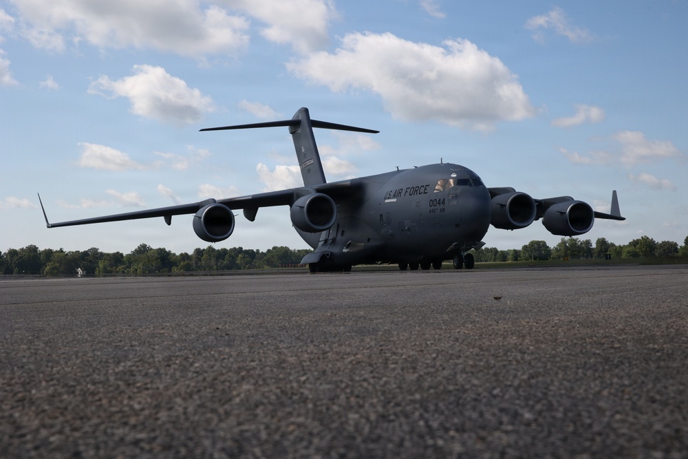 A U.S. Air Force C-17 Globemaster III military cargo aircraft is taxiing or stationary on a tarmac. The aircraft bears the tail number 0044 and is photographed from a low ground-level angle, emphasizing its large size. The setting appears to be a military airfield surrounded by trees under a partly cloudy sky.