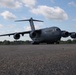 A U.S. Air Force C-17 Globemaster III military cargo aircraft is taxiing or stationary on a tarmac. The aircraft bears the tail number 0044 and is photographed from a low ground-level angle, emphasizing its large size. The setting appears to be a military airfield surrounded by trees under a partly cloudy sky.