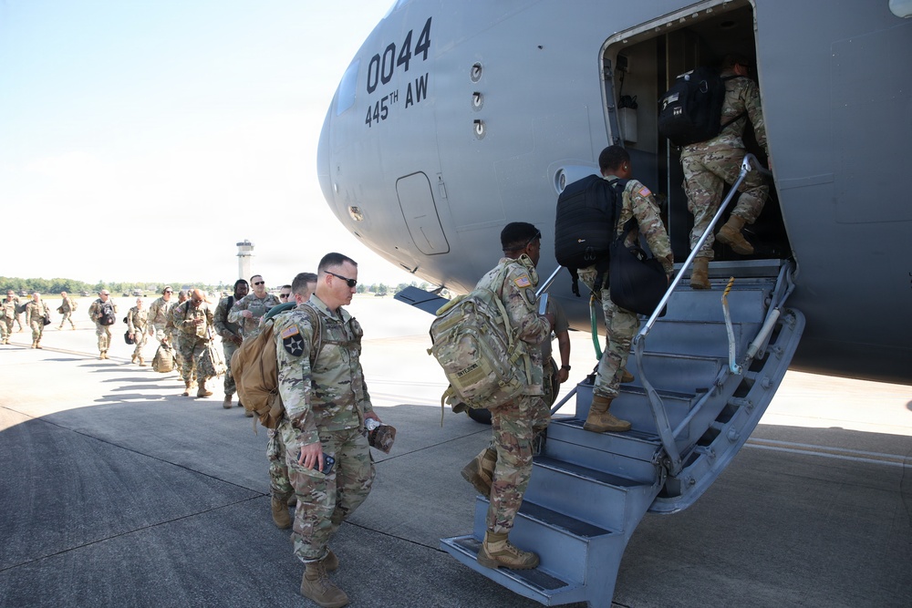 Military personnel in OCP (Operational Camouflage Pattern) uniforms are boarding a large military transport aircraft marked '0044' belonging to the 445th Airlift Wing (445th AW). Soldiers carrying rucksacks and personal gear are queued on the tarmac and ascending the boarding stairs into the aircraft. The scene appears to be a deployment or troop movement operation at a military airfield.