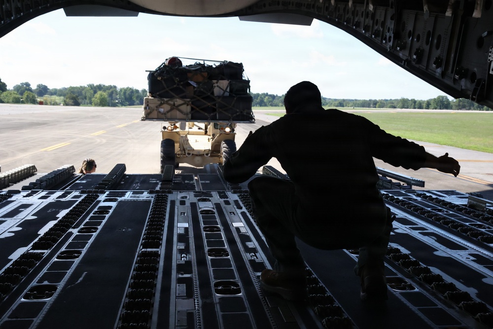 A military loadmaster or cargo handler is crouching on the rear ramp of a large military transport aircraft, directing a forklift carrying a netted cargo pallet toward the aircraft for loading. The scene takes place on a military airfield tarmac surrounded by trees under clear skies. The perspective is from inside the aircraft looking outward toward the ramp opening.