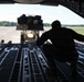 A military loadmaster or cargo handler is crouching on the rear ramp of a large military transport aircraft, directing a forklift carrying a netted cargo pallet toward the aircraft for loading. The scene takes place on a military airfield tarmac surrounded by trees under clear skies. The perspective is from inside the aircraft looking outward toward the ramp opening.