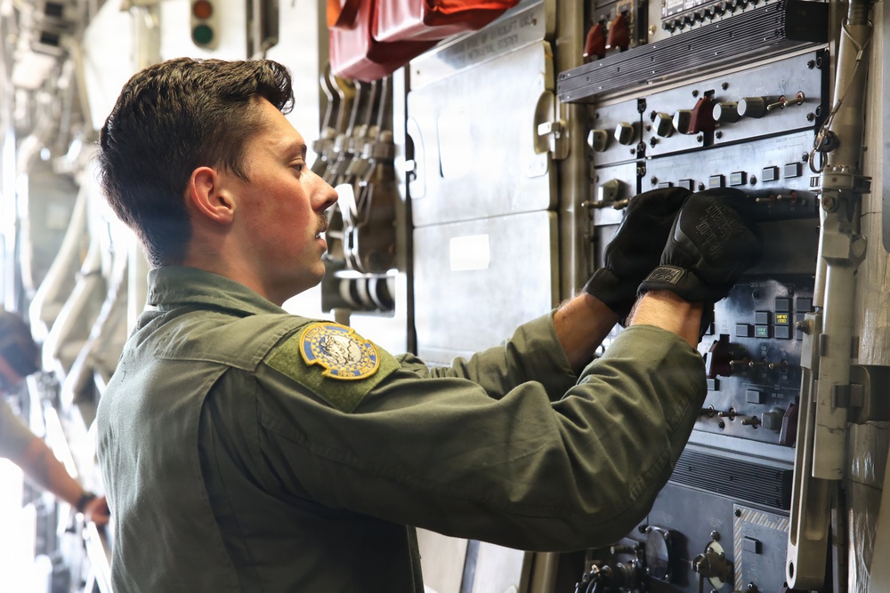 A military service member in an olive green flight suit is operating or inspecting a complex control panel inside what appears to be a military aircraft. The individual is wearing black tactical gloves and has a unit patch visible on the left shoulder. The setting is the interior of a large military transport or cargo aircraft.
