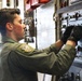 A military service member in an olive green flight suit is operating or inspecting a complex control panel inside what appears to be a military aircraft. The individual is wearing black tactical gloves and has a unit patch visible on the left shoulder. The setting is the interior of a large military transport or cargo aircraft.