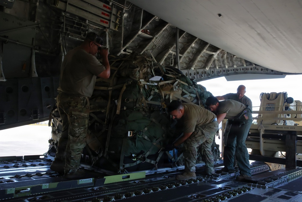 Military personnel are loading or unloading cargo onto a large military transport aircraft, visible from the rear cargo ramp. The cargo appears to consist of bundled equipment secured with straps and rigging, consistent with airdrop or airlift operations. Four service members in military attire are actively handling the heavy cargo bundle on the aircraft's roller-equipped cargo floor.
