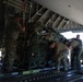 Military personnel are loading or unloading cargo onto a large military transport aircraft, visible from the rear cargo ramp. The cargo appears to consist of bundled equipment secured with straps and rigging, consistent with airdrop or airlift operations. Four service members in military attire are actively handling the heavy cargo bundle on the aircraft's roller-equipped cargo floor.
