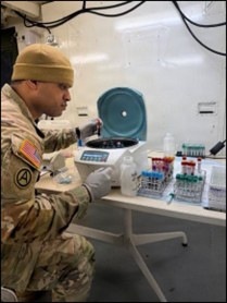 A U.S. Army soldier in camouflage uniform is working in a military medical laboratory setting. The soldier is operating or loading a centrifuge while surrounded by multiple racks of blood collection tubes and laboratory supplies. The setting appears to be a field or forward operating laboratory environment.