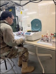 A person wearing military camouflage uniform and tan combat boots is seated at a laboratory workbench, handling blood collection tubes. The setting appears to be a mobile or field medical laboratory, indicated by the compact equipment and utilitarian interior. Multiple blood specimen tubes in a rack and a centrifuge are visible on the workbench.