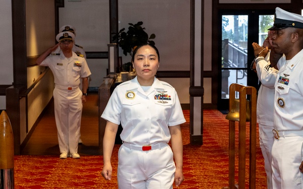 A U.S. Navy military ceremony is taking place in what appears to be a formal venue with red carpeting. A female Navy officer in Service Dress White uniform walks a ceremonial aisle while flanking personnel render salutes on both sides. This appears to be a sideboy honors ceremony, consistent with a change of command, retirement, or promotion event.