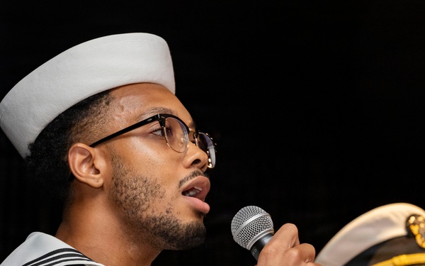 A U.S. Navy enlisted sailor in dress white uniform is speaking into a handheld microphone at what appears to be a formal military ceremony or event. The text 'STC SAN DIEGO' is visible on his uniform collar, indicating an affiliation with a San Diego-based naval command. In the background, a senior naval officer in dress whites with gold-braided cap insignia stands at attention.
