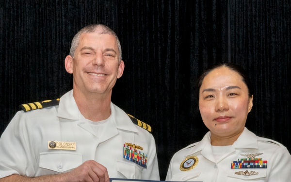 Two U.S. Navy personnel in white dress uniforms are posing together while jointly holding an Honorable Discharge certificate. The officer on the left, identified by his name tag as 'POLONSKY,' appears to hold the rank of Captain (O-6), indicated by four gold stripes on his shoulder boards. The ceremony appears to be a formal military separation or retirement event.