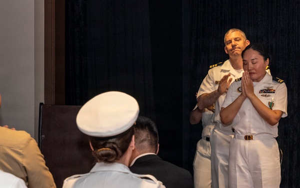 A U.S. Navy military ceremony is taking place in an indoor venue, likely a change of command, retirement, or award ceremony. Two officers in white dress uniforms are on a raised stage, with one appearing to embrace or receive recognition from the other while an audience of uniformed personnel watches. The emotional moment suggests a significant milestone event.