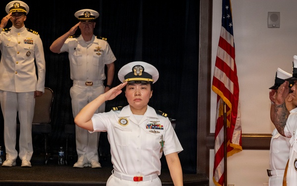 A formal U.S. Navy military ceremony is taking place indoors, likely a change of command or promotion ceremony. A female naval officer in Service Dress White uniform is centered in the frame, rendering a salute while walking between two rows of saluting personnel. Senior officers on a raised stage in the background are also saluting.