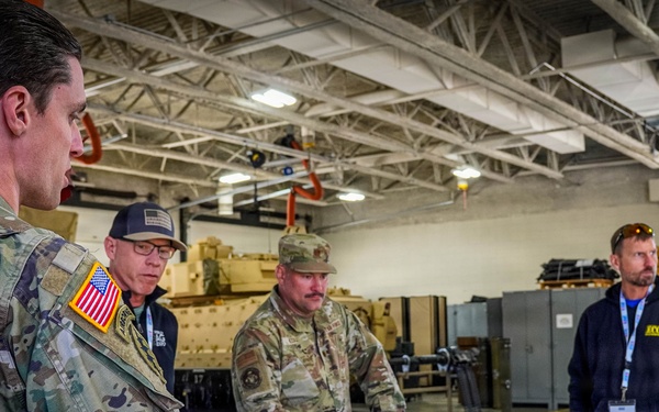 U.S. Army soldiers and civilian observers review tactical map board during military exercise briefing inside vehicle maintenance facility