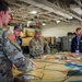 U.S. Army soldiers and civilian observers review tactical map board during military exercise briefing inside vehicle maintenance facility