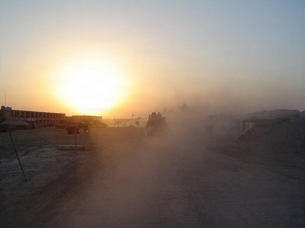 A military convoy moves along a dusty dirt road at either sunrise or sunset in an arid desert environment. Heavy dust clouds obscure visibility, with at least two armored military vehicles visible in silhouette against the low sun. Flat-roofed buildings and earthen berms are visible on both sides of the road, suggesting a forward operating base or military installation.