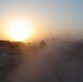 A military convoy moves along a dusty dirt road at either sunrise or sunset in an arid desert environment. Heavy dust clouds obscure visibility, with at least two armored military vehicles visible in silhouette against the low sun. Flat-roofed buildings and earthen berms are visible on both sides of the road, suggesting a forward operating base or military installation.
