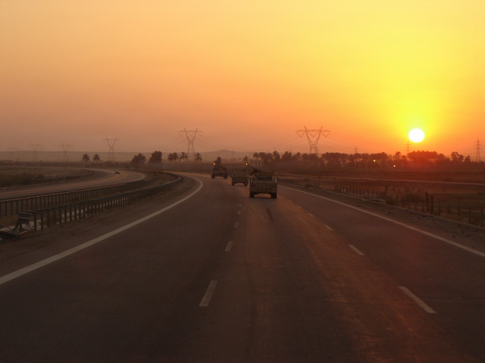 A convoy of military-style vehicles travels along a multi-lane divided highway at sunset or sunrise. The scene is set in an arid, flat landscape with palm trees and high-voltage electrical transmission towers visible in the background. The warm orange and yellow tones of the sun near the horizon dominate the sky.