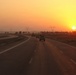 A convoy of military-style vehicles travels along a multi-lane divided highway at sunset or sunrise. The scene is set in an arid, flat landscape with palm trees and high-voltage electrical transmission towers visible in the background. The warm orange and yellow tones of the sun near the horizon dominate the sky.