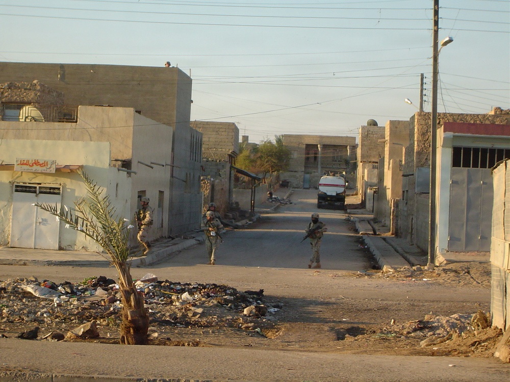 The image shows a military patrol moving through a narrow urban street in what appears to be a Middle Eastern town, based on the architecture and Arabic signage visible on buildings. Three armed soldiers in camouflage uniforms are spread across the street in a tactical formation. The area shows signs of debris and neglect, with a small palm tree in the foreground and utility poles with overhead wires lining the street.