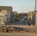The image shows a military patrol moving through a narrow urban street in what appears to be a Middle Eastern town, based on the architecture and Arabic signage visible on buildings. Three armed soldiers in camouflage uniforms are spread across the street in a tactical formation. The area shows signs of debris and neglect, with a small palm tree in the foreground and utility poles with overhead wires lining the street.