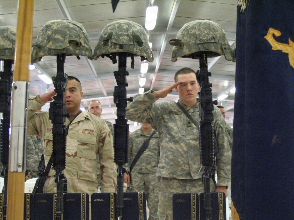 This image depicts a military memorial ceremony, commonly known as a 'Fallen Soldier Battle Cross' or 'Battle Cross Ceremony.' Three rifles are planted bayonet-down with combat helmets placed on top, serving as a traditional tribute to fallen soldiers. U.S. Army personnel in various uniforms are rendering salutes in honor of the deceased.