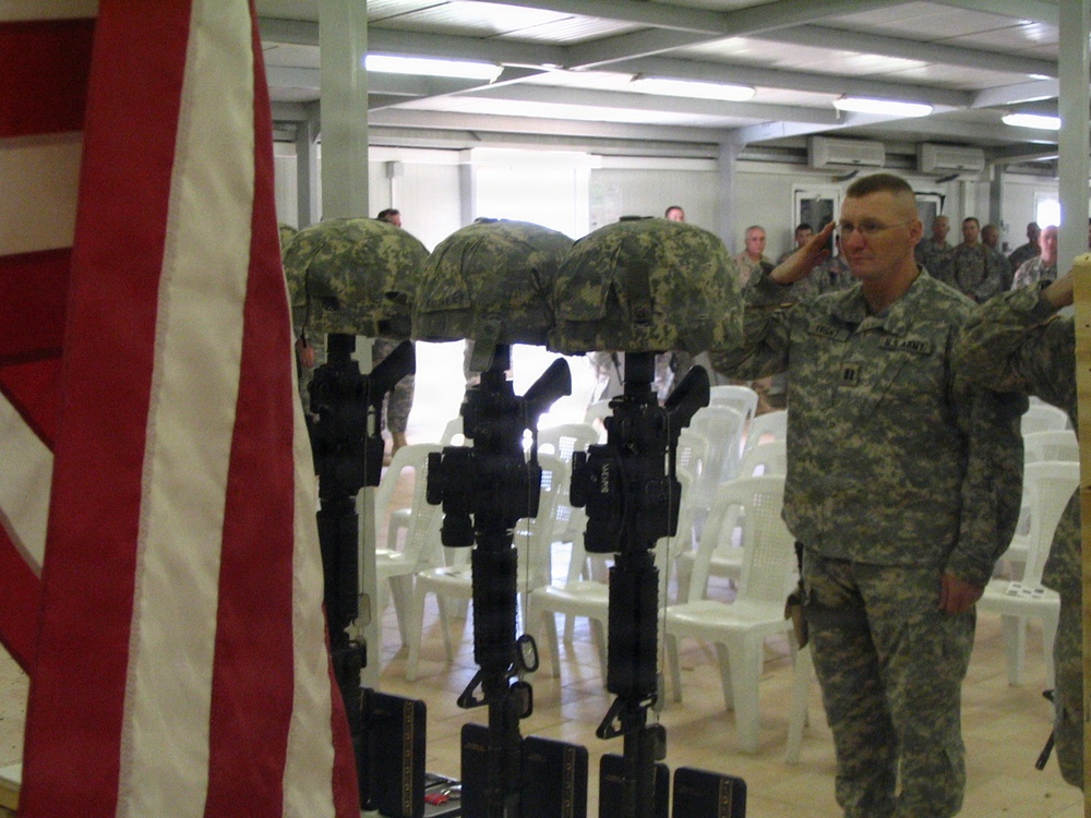 A military memorial ceremony is taking place indoors, likely on a forward operating base. Several 'Battlefield Cross' memorials are displayed — rifles with bayonets fixed into the ground, topped with helmets — honoring fallen soldiers. A U.S. Army officer in ACU (Army Combat Uniform) is rendering a salute alongside other personnel.