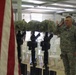 A military memorial ceremony is taking place indoors, likely on a forward operating base. Several 'Battlefield Cross' memorials are displayed — rifles with bayonets fixed into the ground, topped with helmets — honoring fallen soldiers. A U.S. Army officer in ACU (Army Combat Uniform) is rendering a salute alongside other personnel.