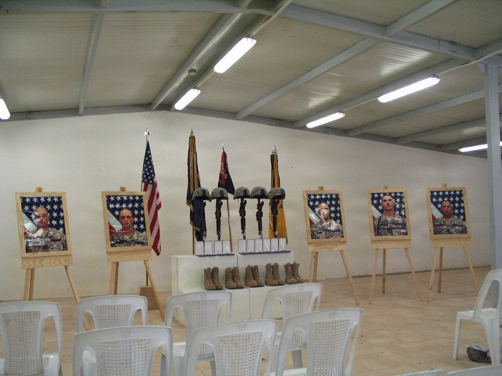 This image depicts a military memorial ceremony setup inside a prefabricated building, honoring fallen soldiers. Six portrait photographs of service members in uniform are displayed on wooden easels, each backed by American flags. In the center, combat boots, rifles with bayonets fixed into the ground, helmets, and dog tags are arranged in the traditional 'Battlefield Cross' memorial formation.