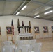 This image depicts a military memorial ceremony setup inside a prefabricated building, honoring fallen soldiers. Six portrait photographs of service members in uniform are displayed on wooden easels, each backed by American flags. In the center, combat boots, rifles with bayonets fixed into the ground, helmets, and dog tags are arranged in the traditional 'Battlefield Cross' memorial formation.