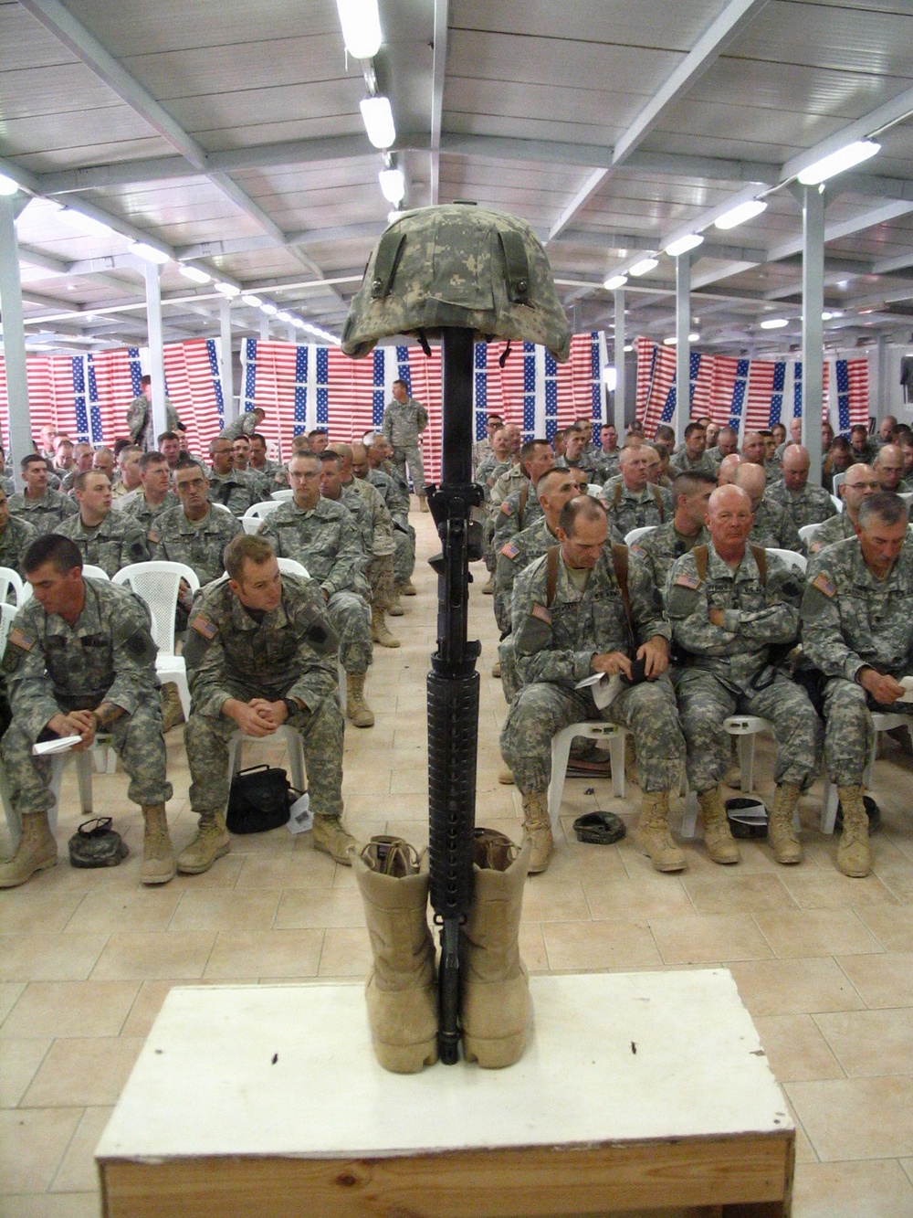 A military memorial ceremony is taking place inside a large indoor facility, likely on a forward operating base. A 'Battlefield Cross' — consisting of a rifle with bayonet fixed into boots, topped with a combat helmet — is displayed on a platform in the foreground. Hundreds of soldiers in U.S. Army ACU (Army Combat Uniform) are seated in rows facing the memorial, with American flags decorating the walls in the background.