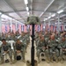 A military memorial ceremony is taking place inside a large indoor facility, likely on a forward operating base. A 'Battlefield Cross' — consisting of a rifle with bayonet fixed into boots, topped with a combat helmet — is displayed on a platform in the foreground. Hundreds of soldiers in U.S. Army ACU (Army Combat Uniform) are seated in rows facing the memorial, with American flags decorating the walls in the background.