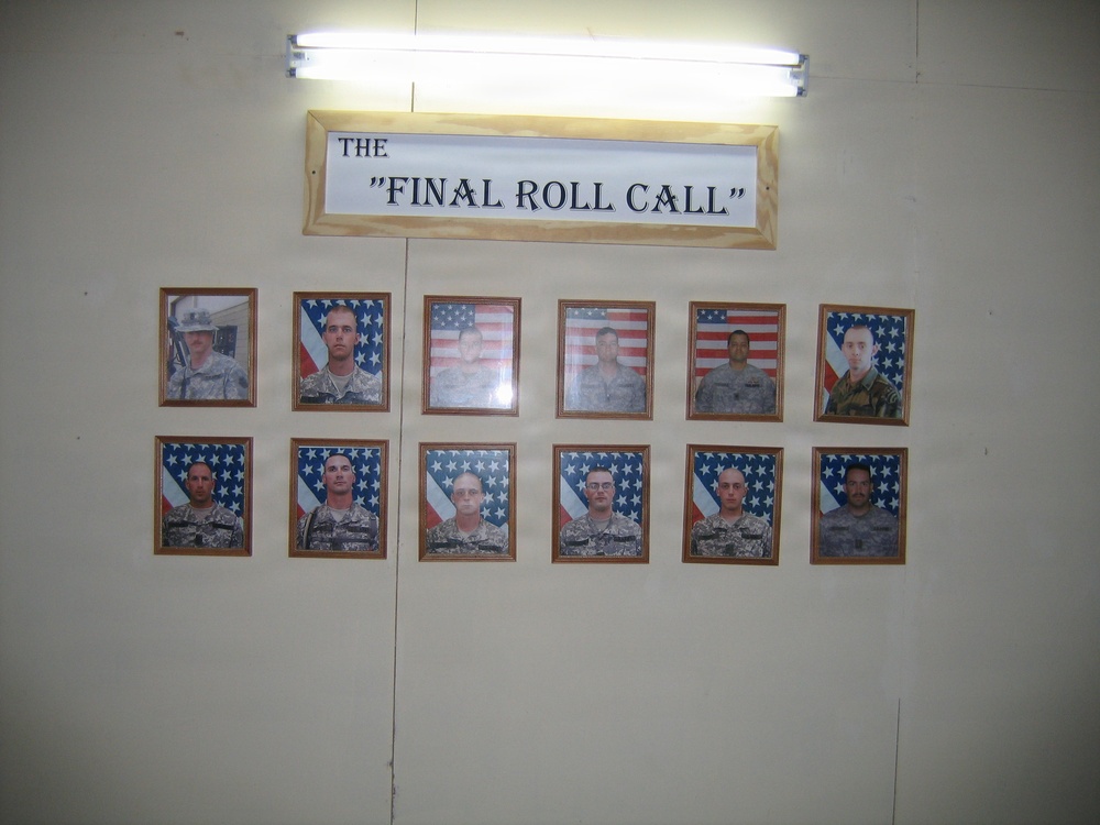 A memorial wall display titled 'The Final Roll Call' is mounted on a light-colored wall, illuminated by a fluorescent light above. The display features twelve framed photographs of military personnel arranged in two rows of six, most photographed against American flag backgrounds. This type of display is a traditional military tribute honoring fallen service members.